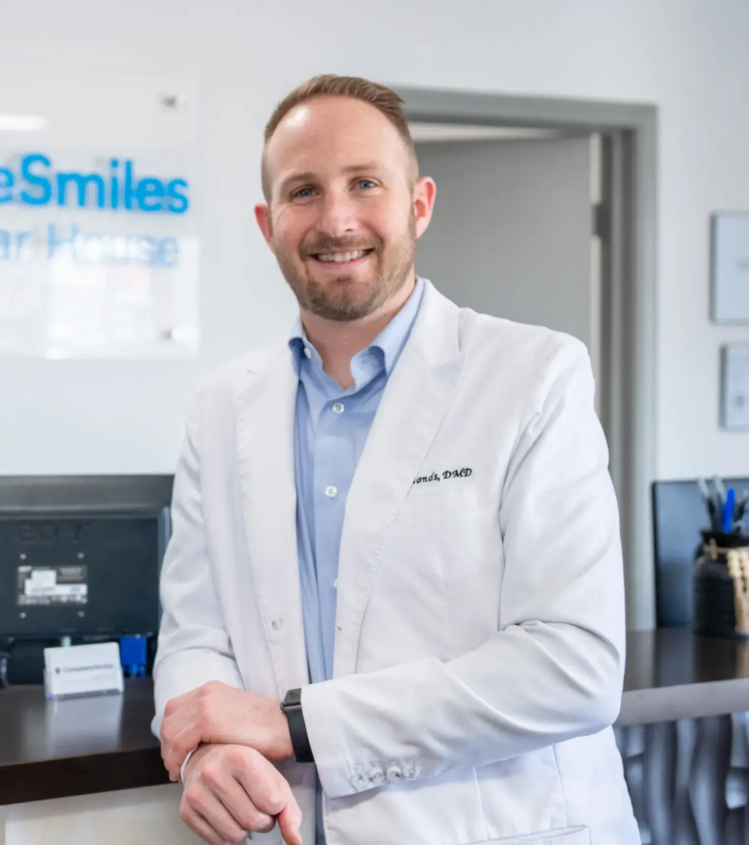 Smiling male dentist wearing white coat standing in a dental office reception area.