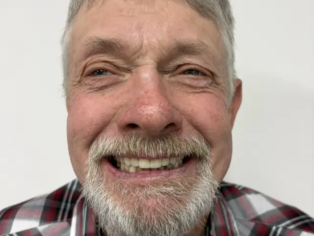 Close-up of an older man with gray hair and beard smiling broadly, wearing a red and black plaid shirt.