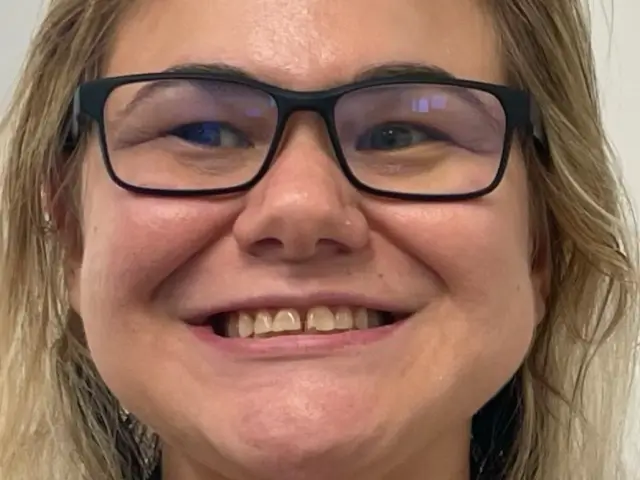 Close-up of a smiling woman with light skin, blonde hair, and black glasses.