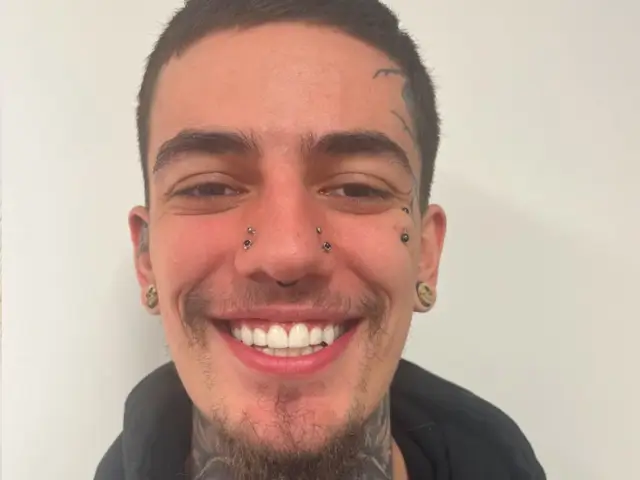 Smiling young man with multiple facial piercings, ear gauges, and neck tattoos wearing a black hoodie.
