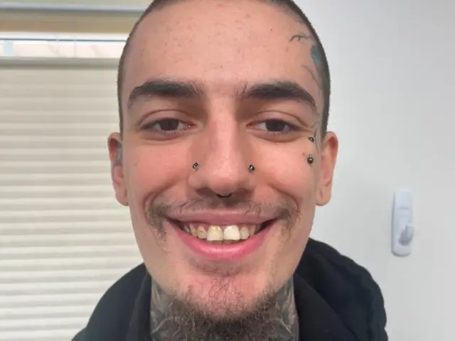 Close-up of a smiling man with several facial piercings and tattoos, wearing a black hoodie.