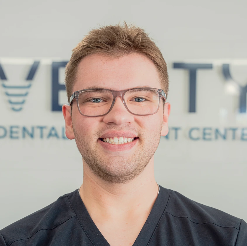 Smiling young man with glasses and short hair wearing a black medical scrub top in front of a dental clinic sign.