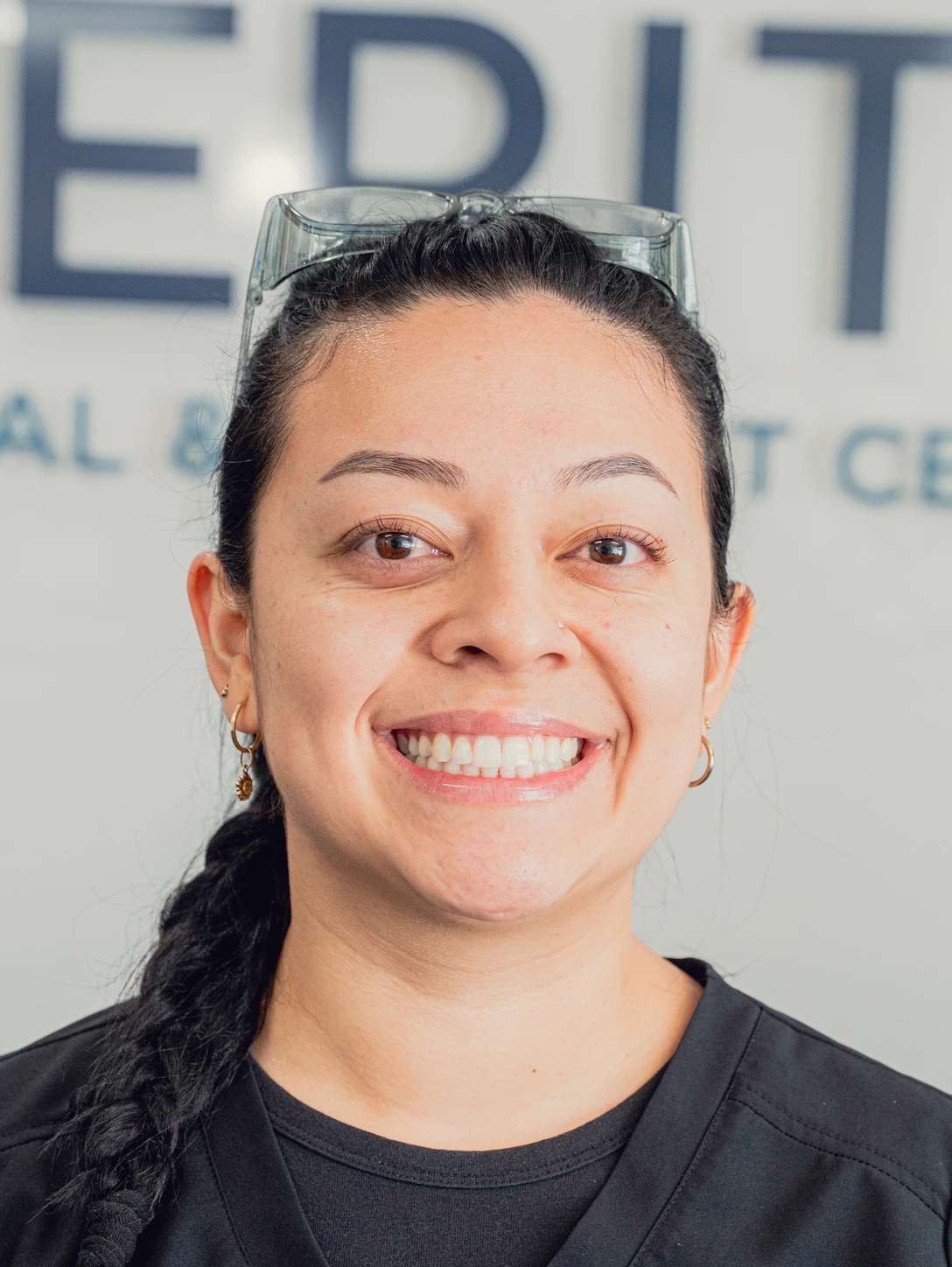 Smiling woman with braided hair, wearing black scrubs and safety glasses on her head.