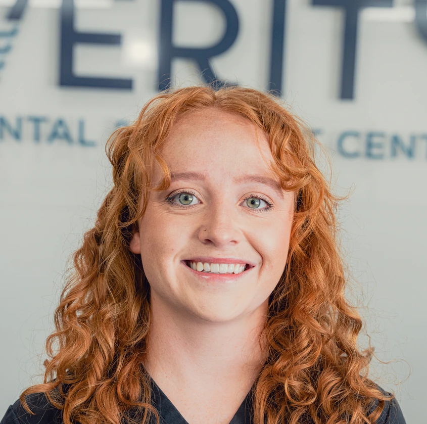 Smiling young woman with curly red hair and green eyes wearing a black top against a blurred light background with dark blue text.