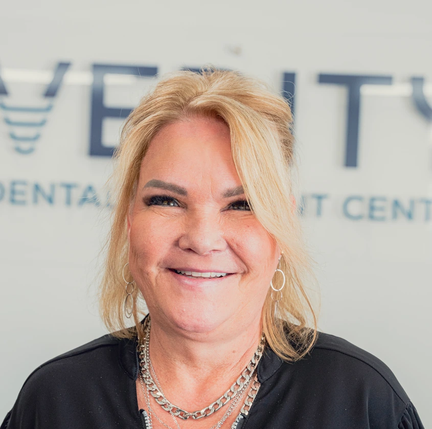 Smiling middle-aged woman with blonde hair wearing hoop earrings and layered silver necklaces, standing in front of a dental clinic sign.