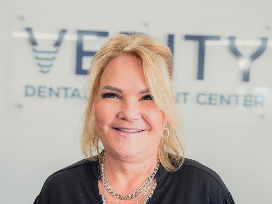 Smiling middle-aged woman with blonde hair wearing silver hoop earrings and chains, standing in front of a dental implant center sign.