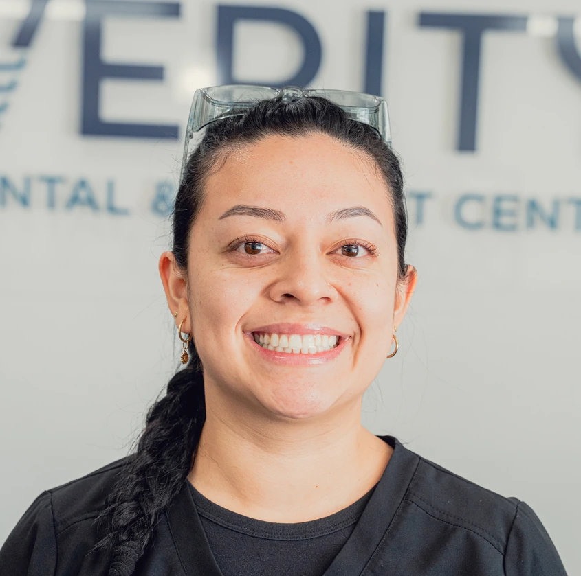 Smiling woman with braided hair and protective glasses on her head wearing black scrubs in front of a dental clinic sign.
