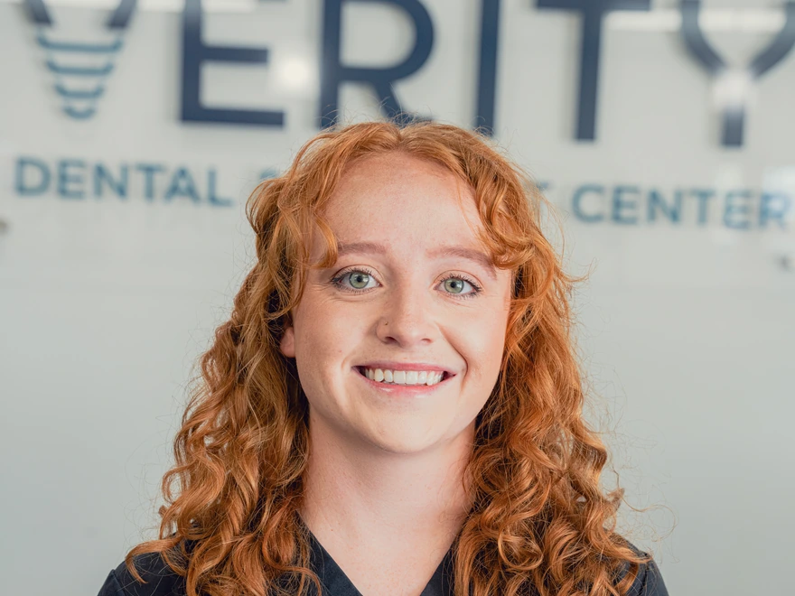 Smiling woman with curly red hair standing in front of a dental center sign.