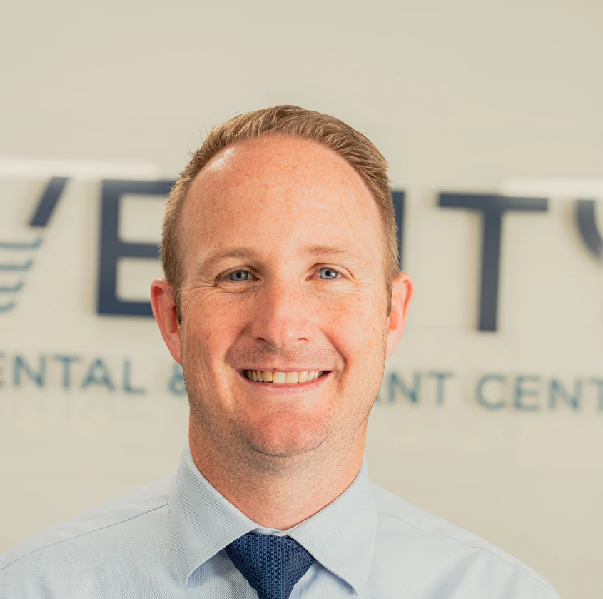 Smiling man with short light brown hair wearing a light blue shirt and navy tie in front of a dental office sign.