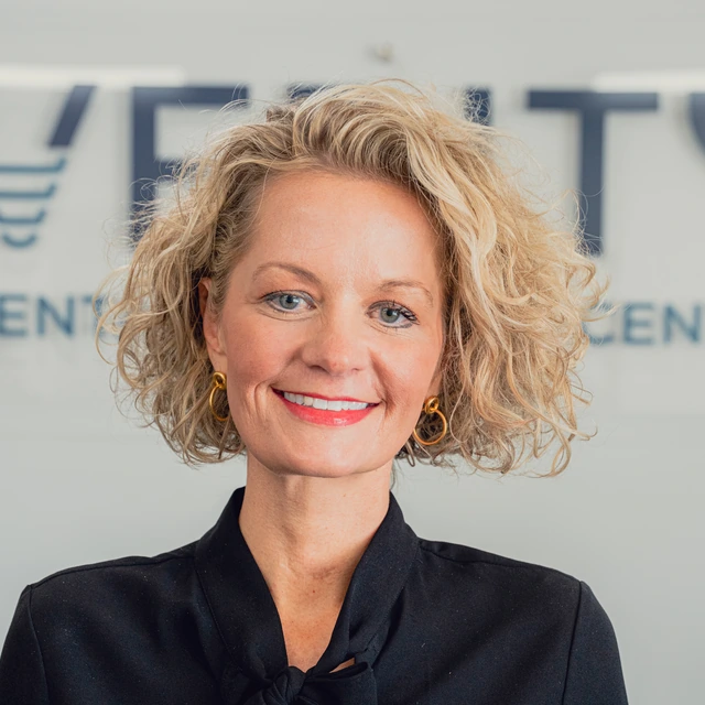 Smiling woman with curly blonde hair wearing gold earrings and a black blazer, standing in front of a blurred office sign.