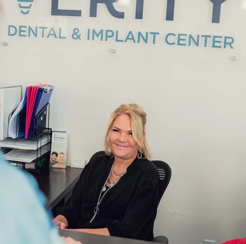 Smiling woman with blonde hair sitting at a dental reception desk with 'Dental & Implant Center' sign in the background.