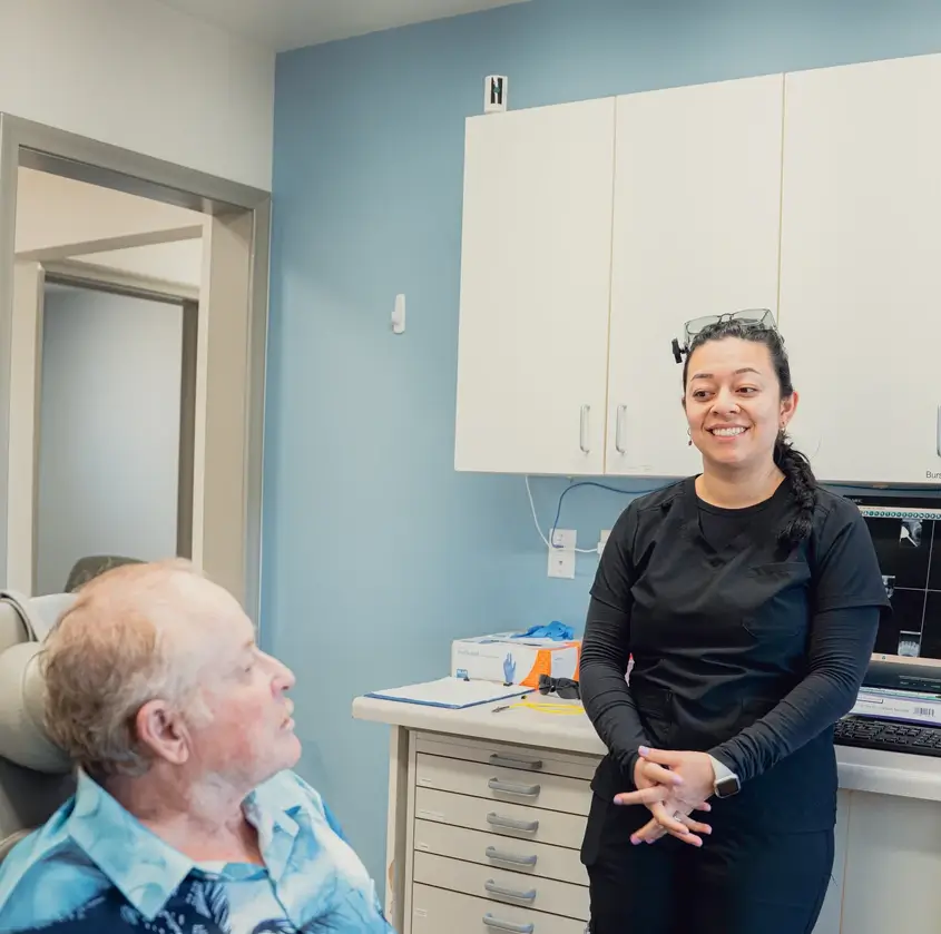 Smiling female healthcare professional speaking with an elderly male patient seated in a medical office.