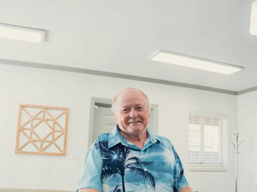 Smiling elderly man wearing a blue Hawaiian shirt with palm tree patterns, sitting indoors with white walls and a wooden geometric decoration in the background.