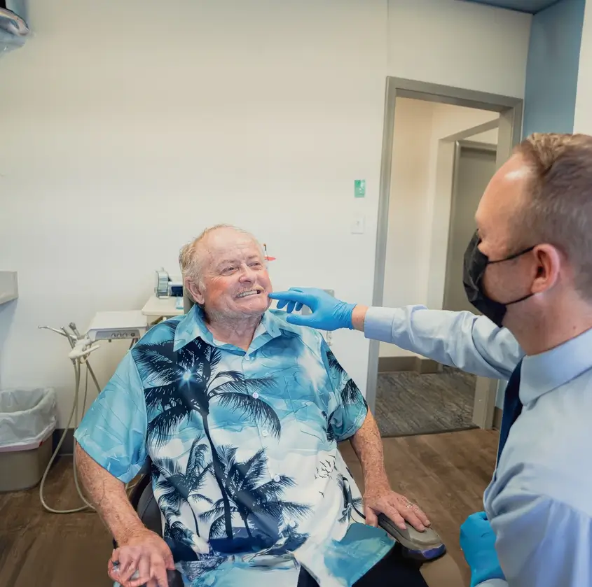 Dentist wearing gloves and mask examining the teeth of an elderly man in a blue Hawaiian shirt sitting in a dental chair.