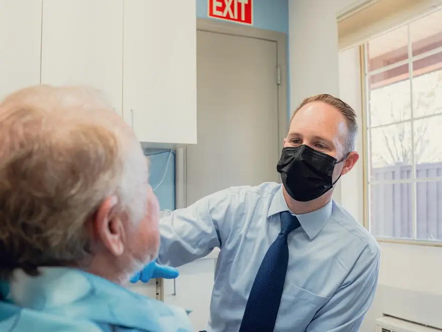 Healthcare professional wearing a black mask and blue shirt with tie attending to an elderly patient in a medical office.