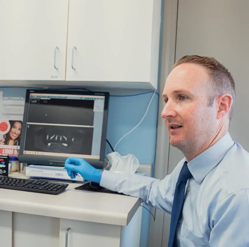 Dentist wearing blue gloves pointing at dental X-ray images on a computer screen in a clinic.