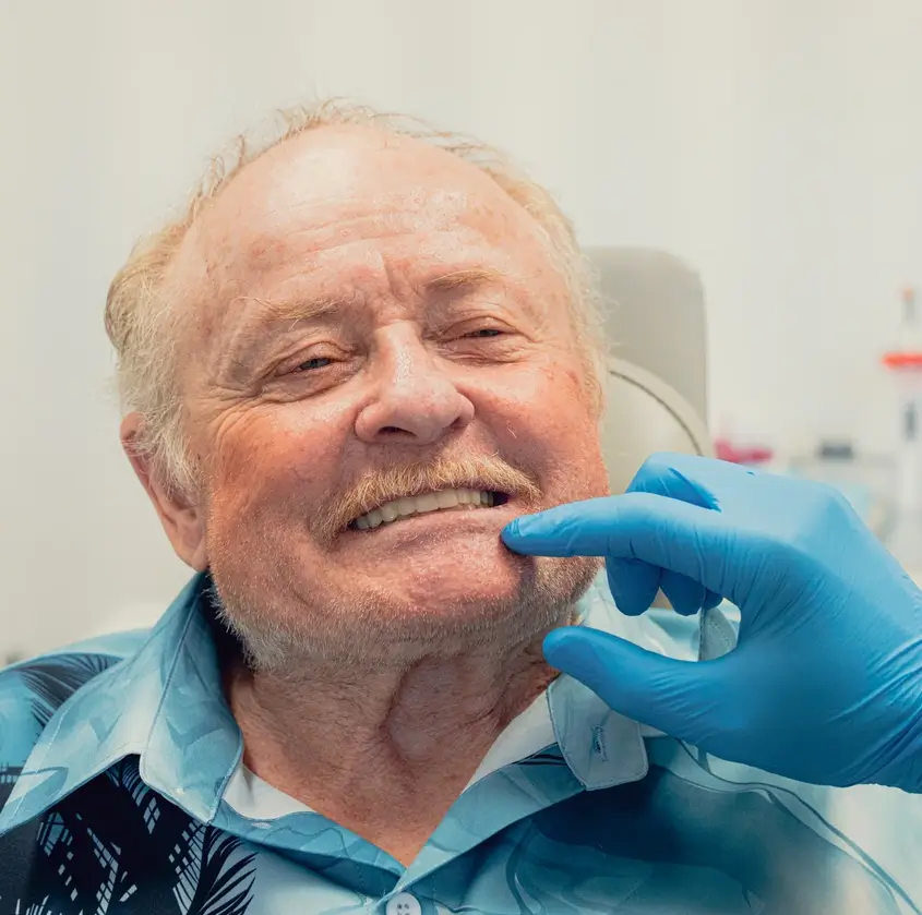 Dentist wearing blue gloves examining an elderly man's teeth as he smiles.