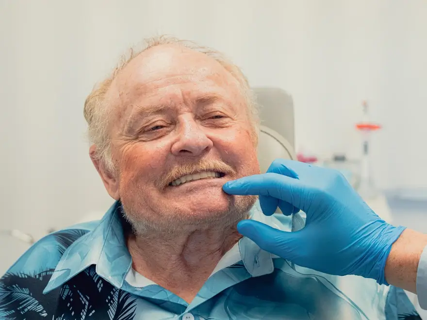 Elderly man smiling gently while a person wearing blue medical gloves examines his lower jaw.