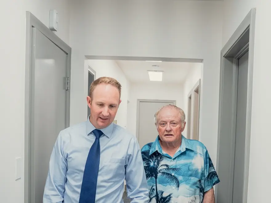 Middle-aged man in a blue shirt and tie walking down a hallway with an elderly man in a blue palm tree print shirt.