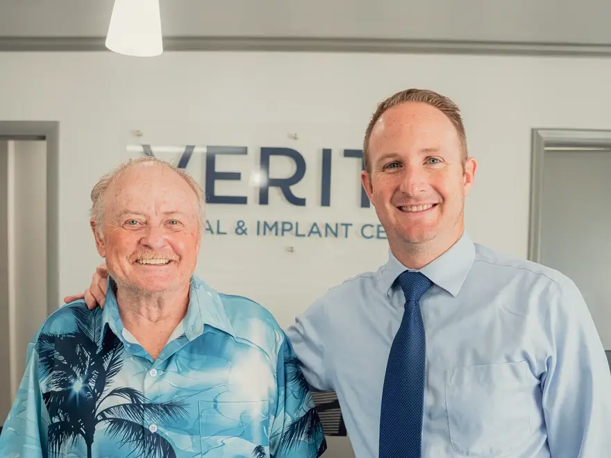 Smiling older man in a blue Hawaiian shirt and a younger man in a blue shirt and tie standing together in front of a dental and implant center sign.