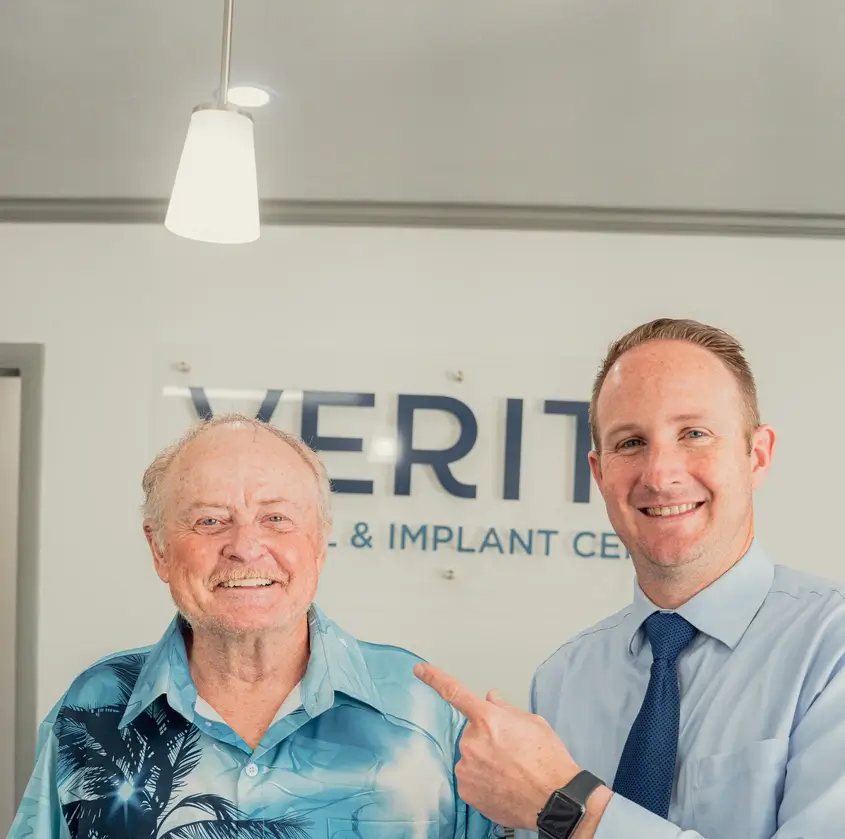 Smiling older man in a blue tropical shirt standing next to a younger man in a light blue shirt and navy tie who is pointing at him, both in a dental clinic.