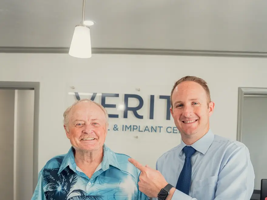 Smiling doctor in a blue shirt and tie pointing at an older man in a blue palm tree shirt inside a clinic.