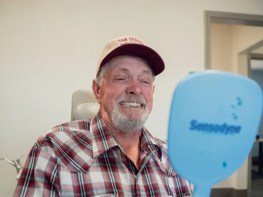 Older man with a gray beard smiling and looking at a blue Sensodyne mirror, wearing a red and white plaid shirt and a cap.