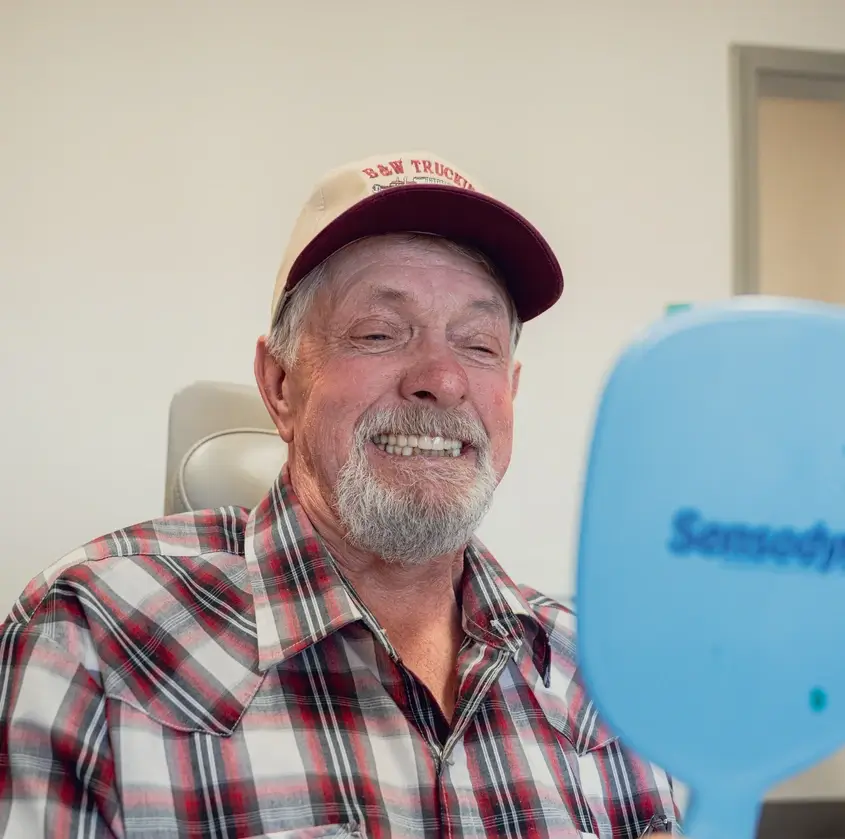Older man with a gray beard and mustache wearing a red and gray plaid shirt and beige cap smiling at a hand mirror.
