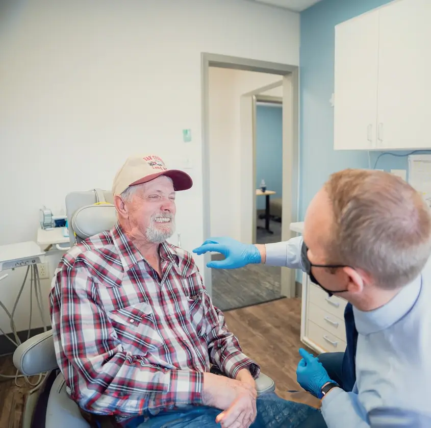 Smiling elderly man in a plaid shirt and cap sits in a dentist chair while a dentist wearing blue gloves talks to him.