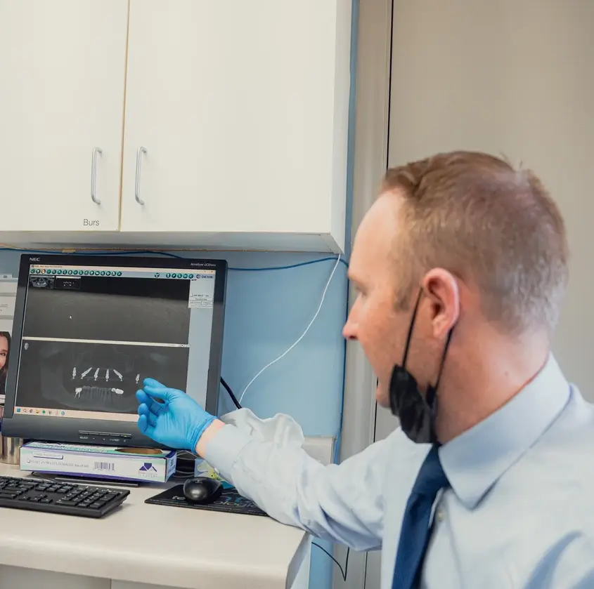 Man wearing blue gloves and a face mask pointing at dental X-ray images on a computer screen in a clinical setting.