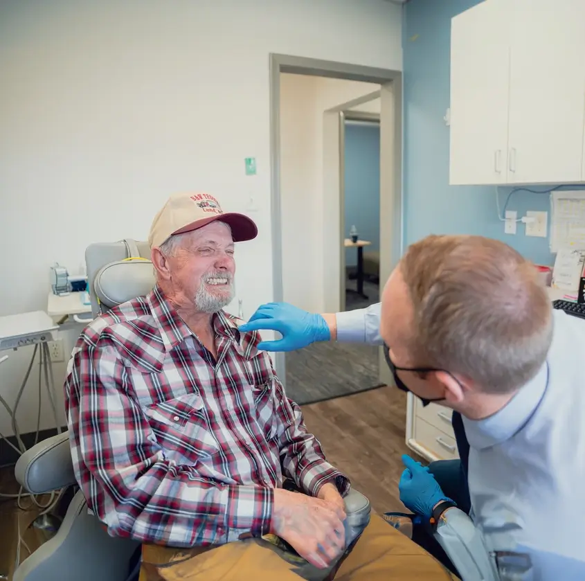 A male healthcare professional wearing blue gloves examines an elderly man in a plaid shirt and cap during a medical checkup in a clinic.