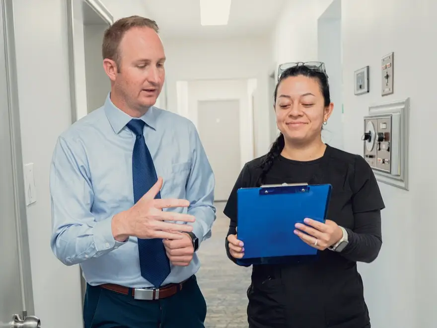 Man in blue dress shirt and tie talking to woman in black scrubs holding a blue clipboard in a hallway.
