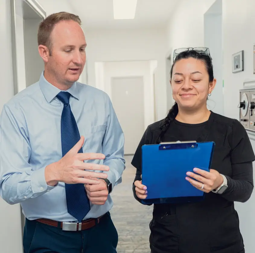 A man in business attire discusses something with a woman in black scrubs holding a blue clipboard in a hallway.