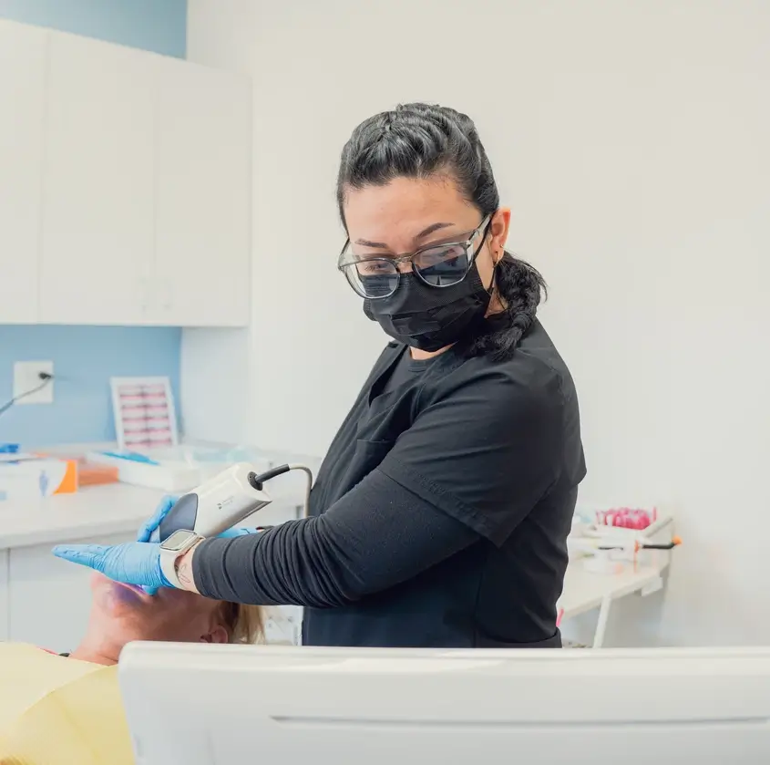 Dental professional wearing black scrubs, gloves, glasses, and a face mask using a handheld dental scanner on a patient in a dental clinic.