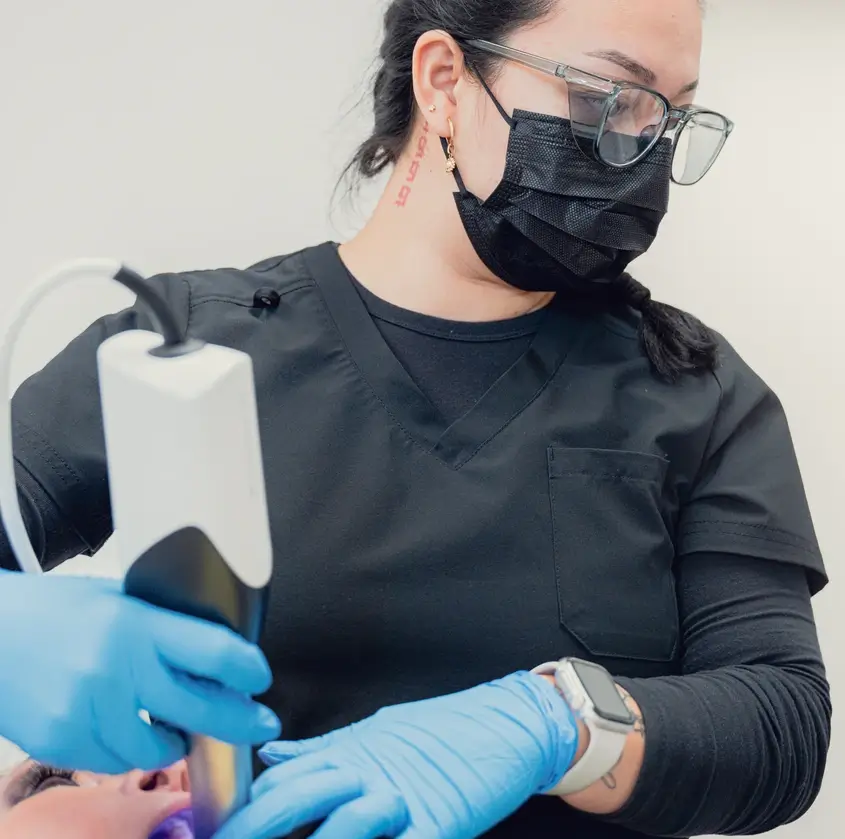 Dental professional wearing black scrubs, a black face mask, and glasses using a dental scanner on a patient.