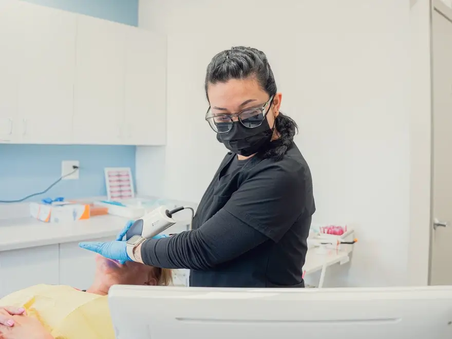A healthcare professional wearing a black mask and gloves performs a facial treatment on a patient lying down in a clinical setting.