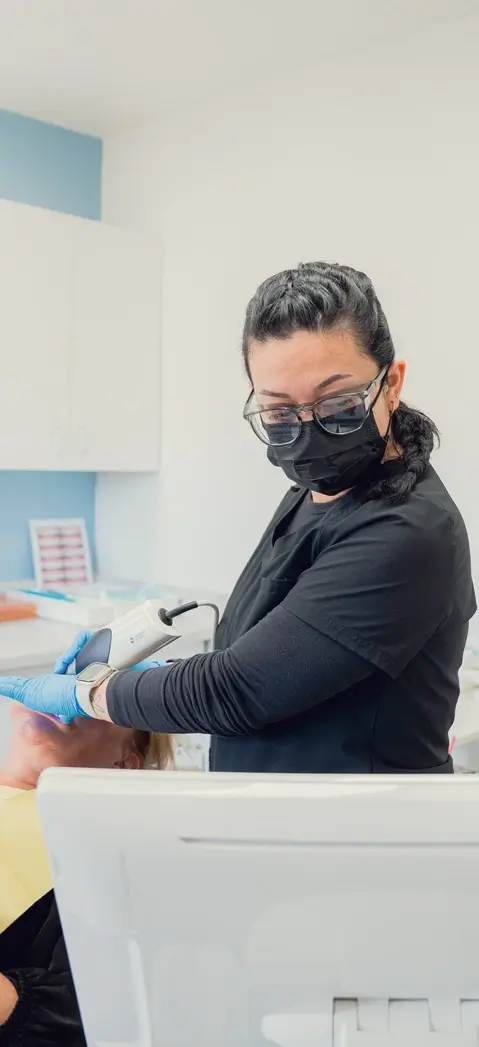 Healthcare professional wearing black scrubs, gloves, glasses, and a face mask operates a handheld medical device on a patient lying back in a clinical setting.