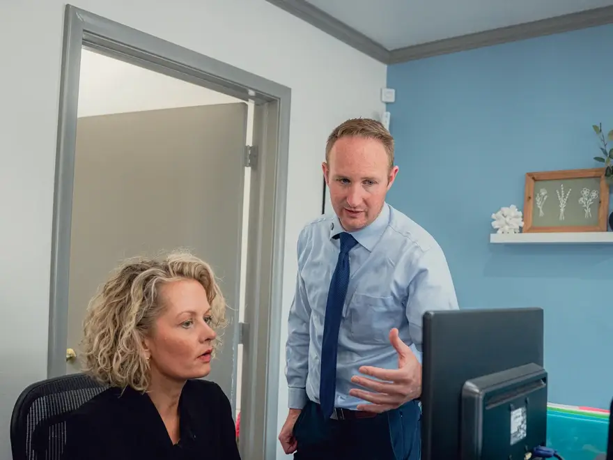 A man in a light blue shirt and dark blue tie explains something to a seated woman with curly blonde hair in an office setting.