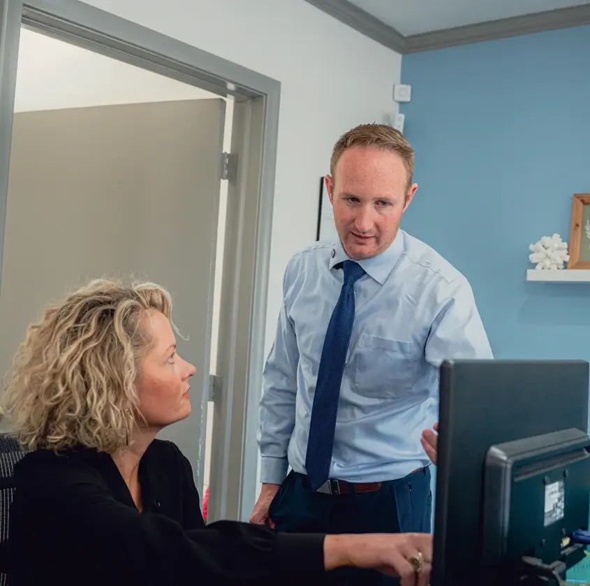 Man in light blue shirt and blue tie standing and talking to seated woman with curly blonde hair near a computer monitor in an office.