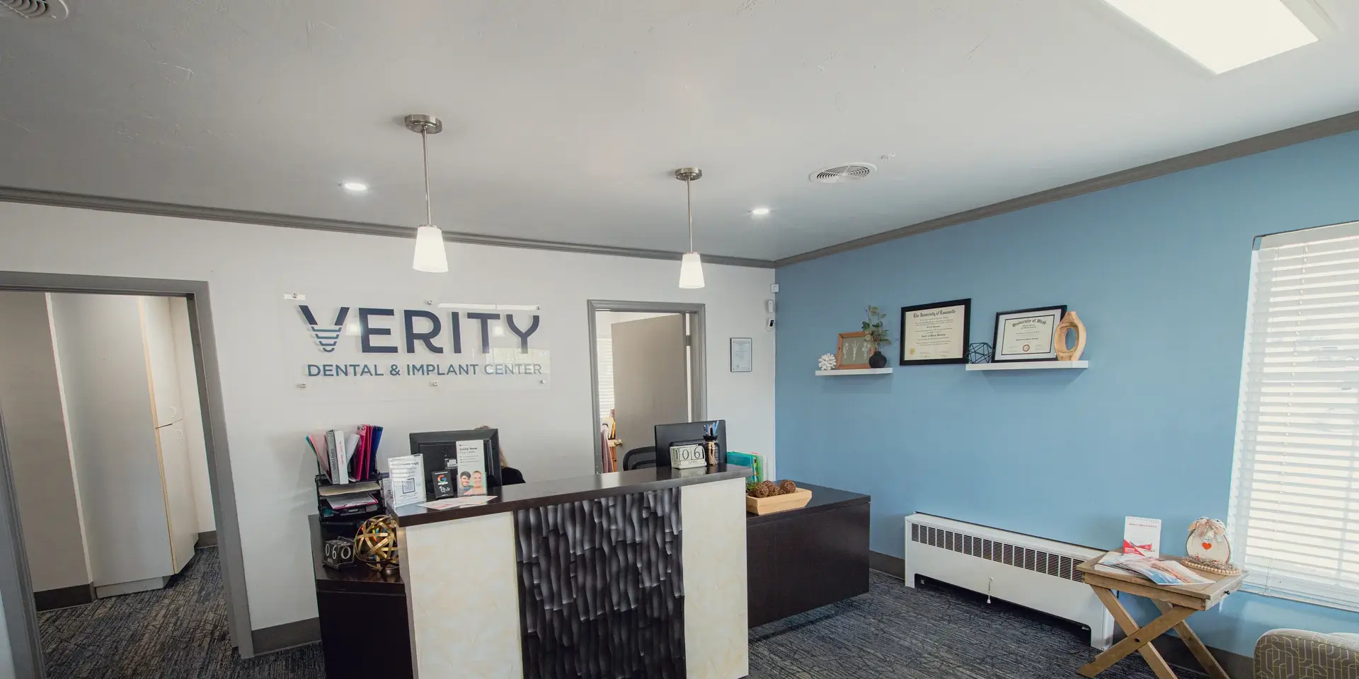 Reception area of Verity Dental & Implant Center with a desk, office supplies, certificates on a blue wall, and natural light from a window.