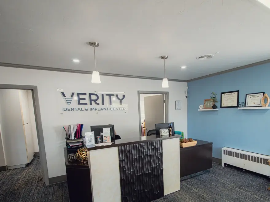 Reception desk area at Verity Dental & Implant Center with chairs, computer monitors, certificates on a blue wall, and two pendant lights.