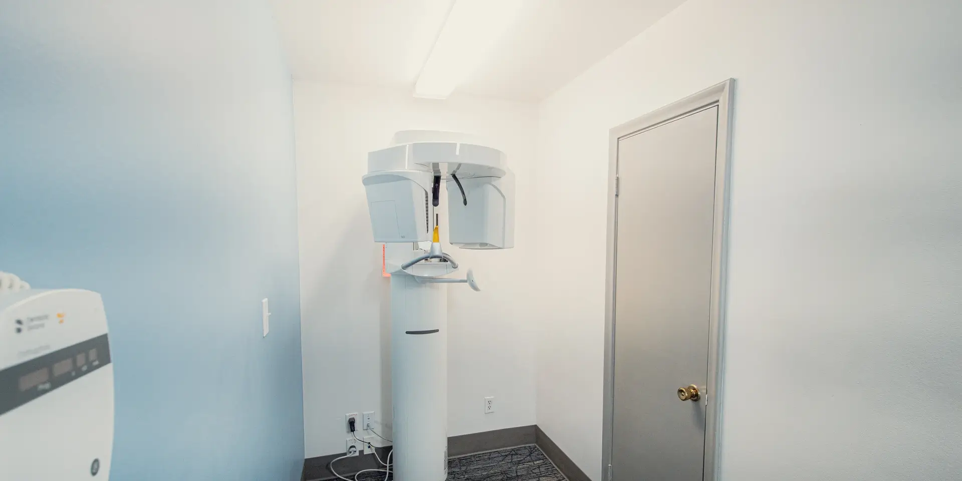 Dental panoramic X-ray machine in a small clinical room with white and blue walls and a closed gray door.