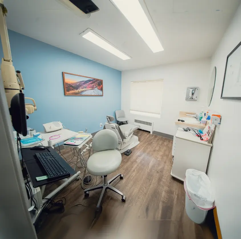 Small dental examination room with blue accent wall, dental chair, stool, desk with keyboard, and medical supplies.
