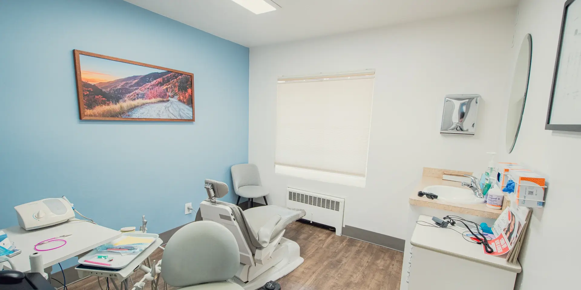 Bright dental examination room with dental chair, tools, sink, and a scenic mountain road photograph on a blue accent wall.