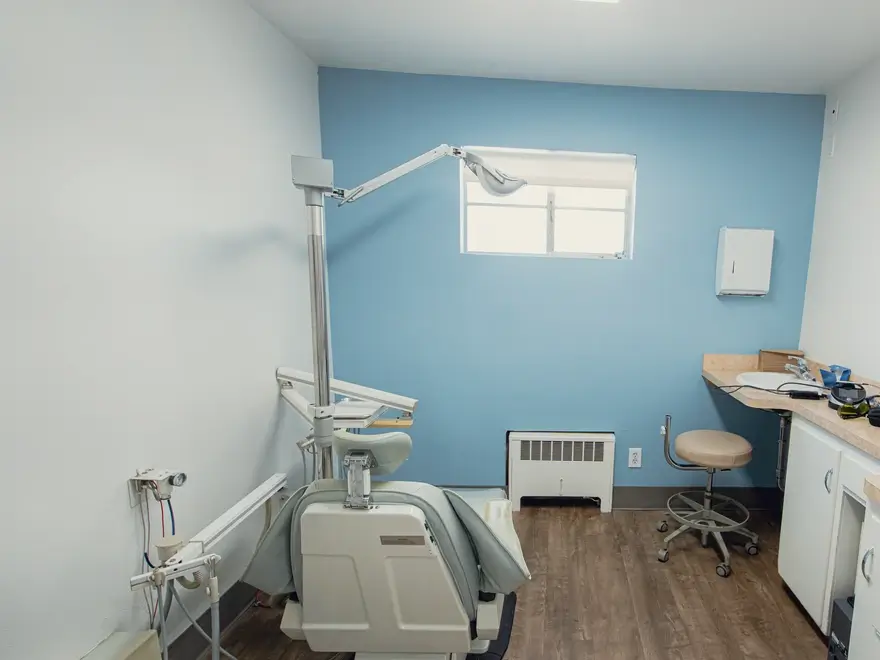 Empty dental examination room with dental chair, overhead light, stool, counter, and sink, featuring blue and white walls and wooden floor.