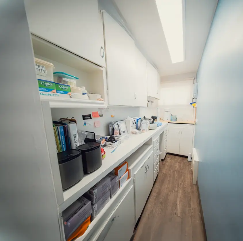 Narrow room with white cabinets and shelves stocked with medical supplies and documents, featuring wooden flooring and pale blue walls.