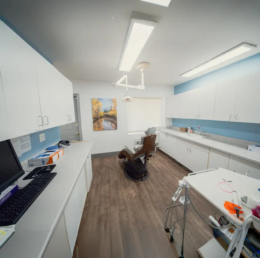 A clean dental office room with a dental chair in the center, white cabinets along the walls, a computer on the counter, and medical tools on a wheeled cart.