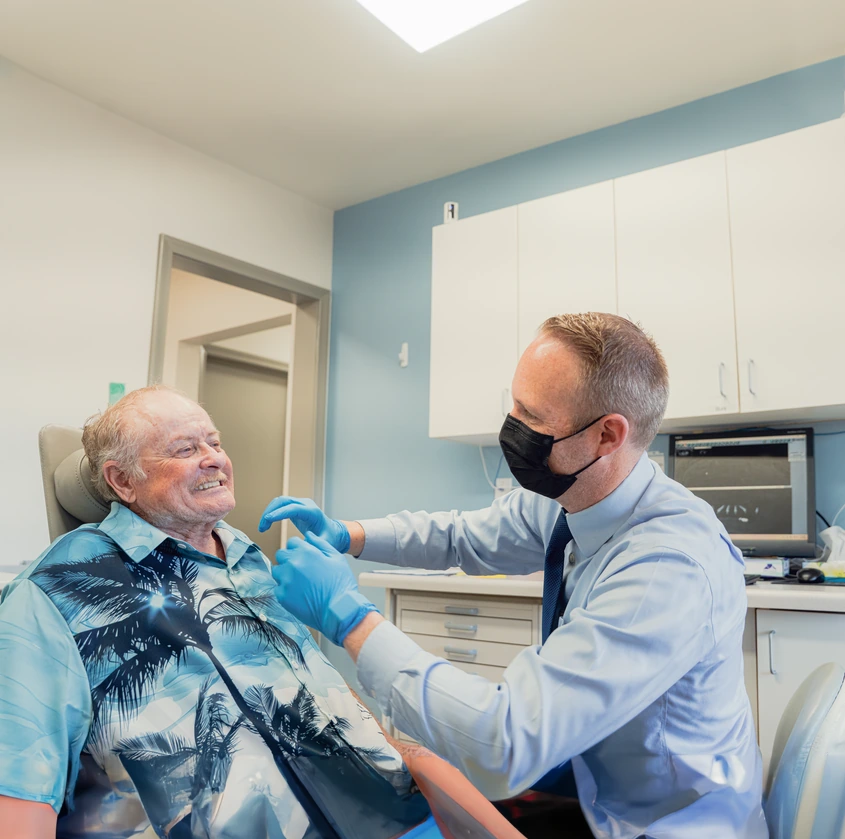 Dentist wearing a mask and gloves examining an elderly male patient in a dental office.