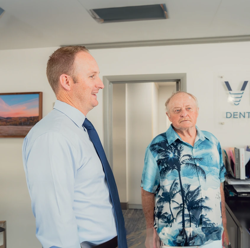 Two men talking in a dental office reception area, one wearing a light blue shirt and tie, the other in a blue and white palm tree print shirt.
