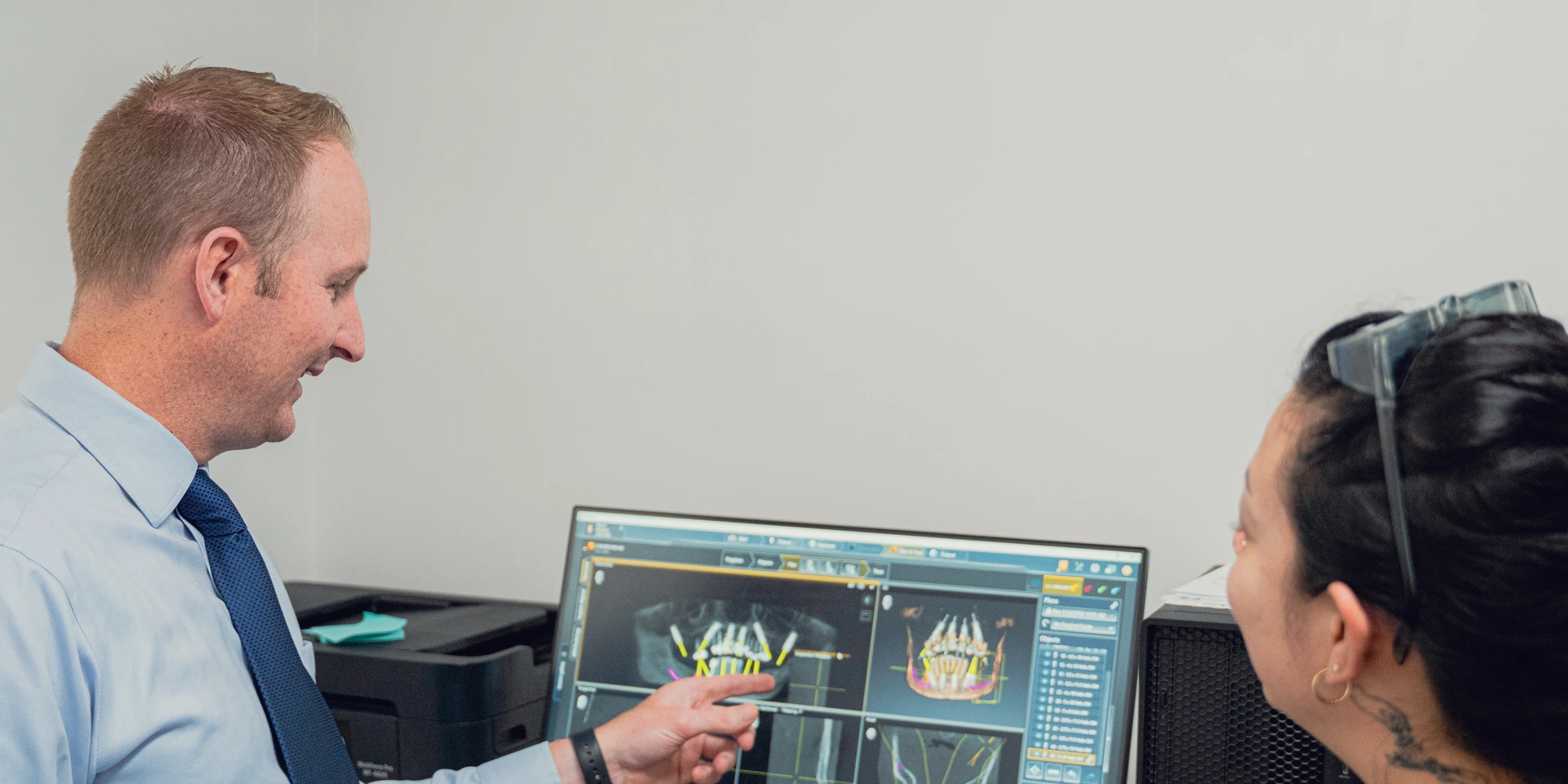Man in blue shirt and tie pointing at dental X-ray images on a computer screen while a woman listens.